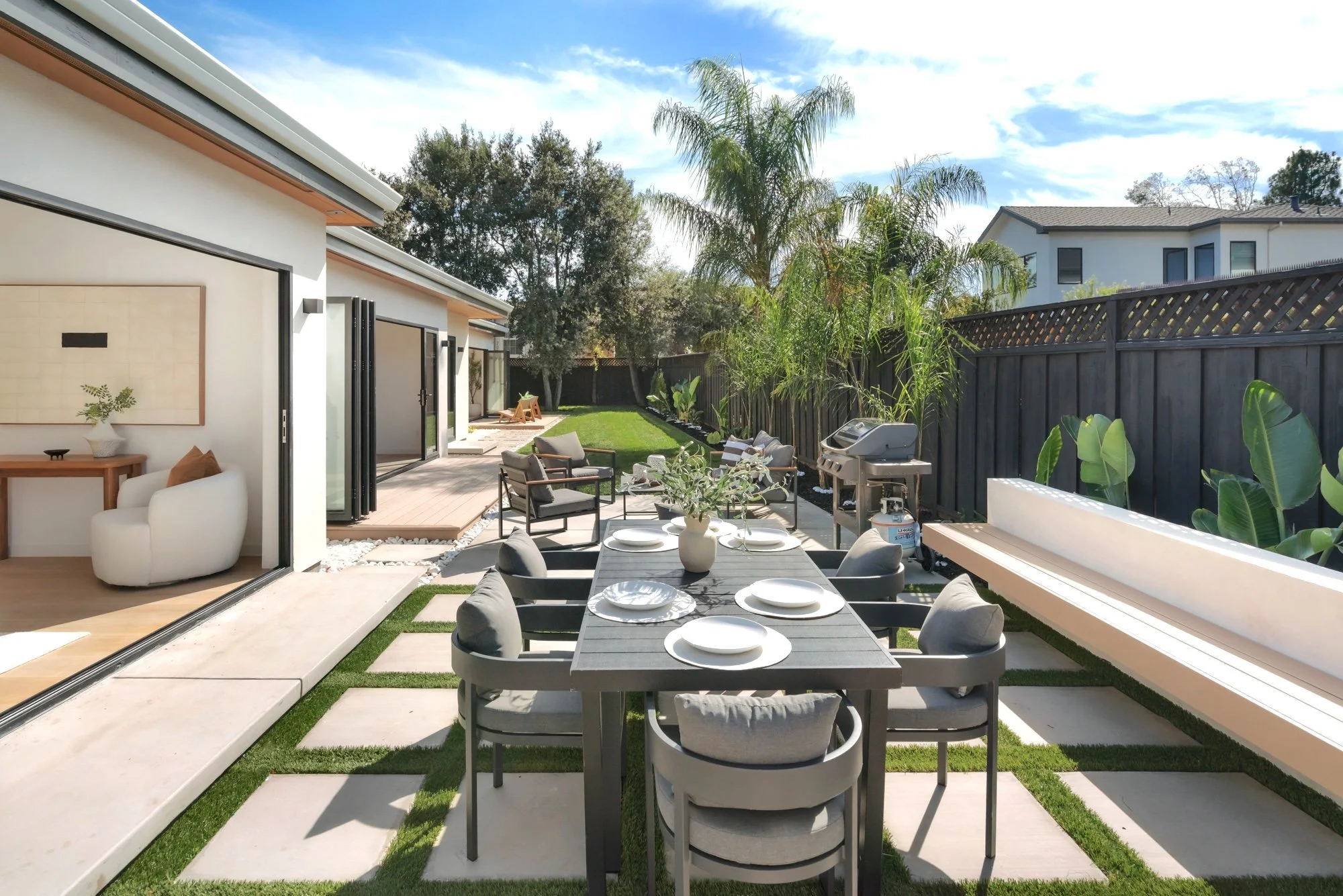 A backyard patio with a dining table set with white plates and cushions on the chairs, outdoor seating, a grill, lush greenery, a wooden fence, and a modern house with large windows, under a partly cloudy sky.