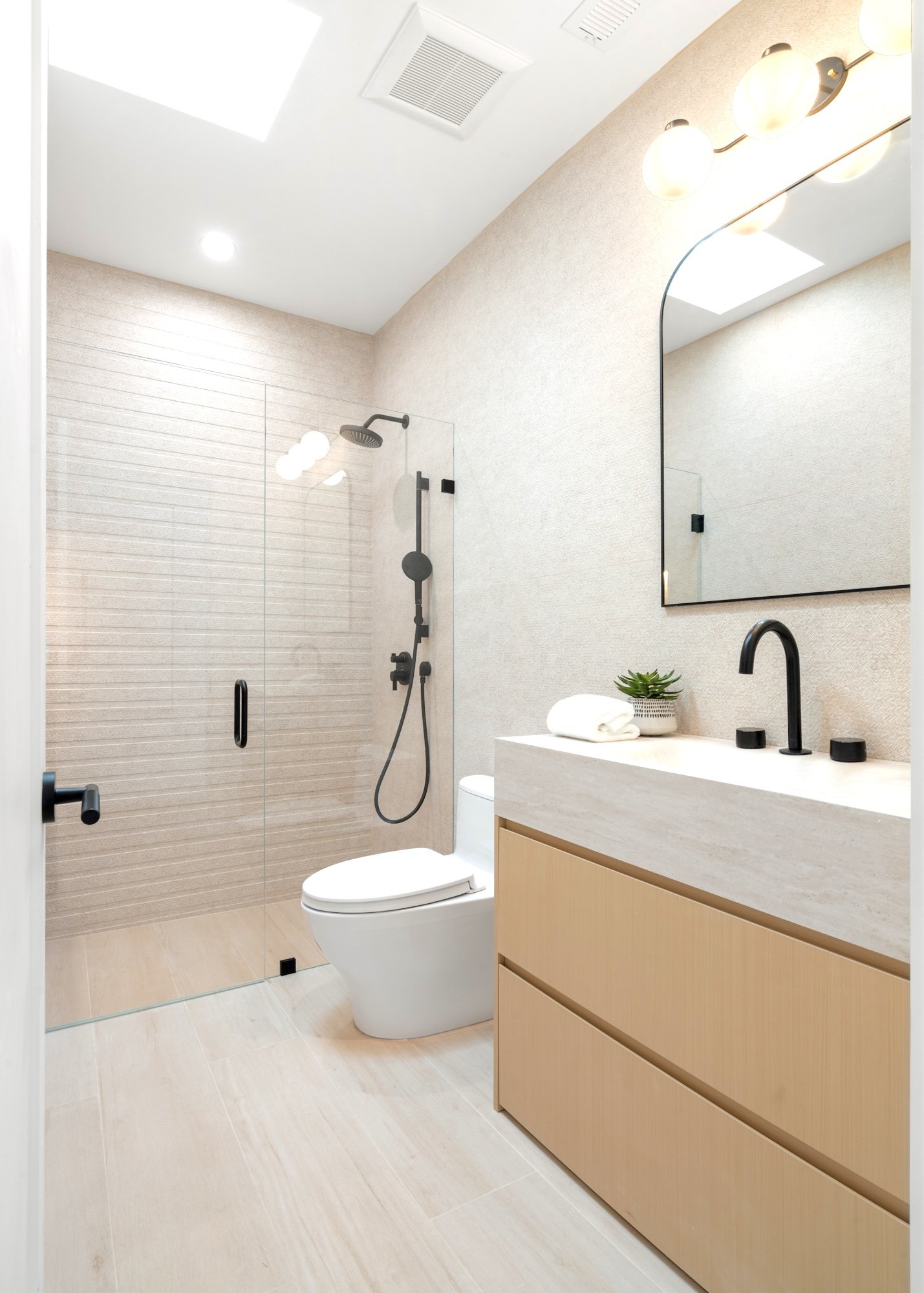A modern bathroom with beige walls and floor, a glass shower door with a black handle, a black showerhead, a white toilet, a beige vanity with a black faucet, a large mirror, a small decorative plant, and a towel.