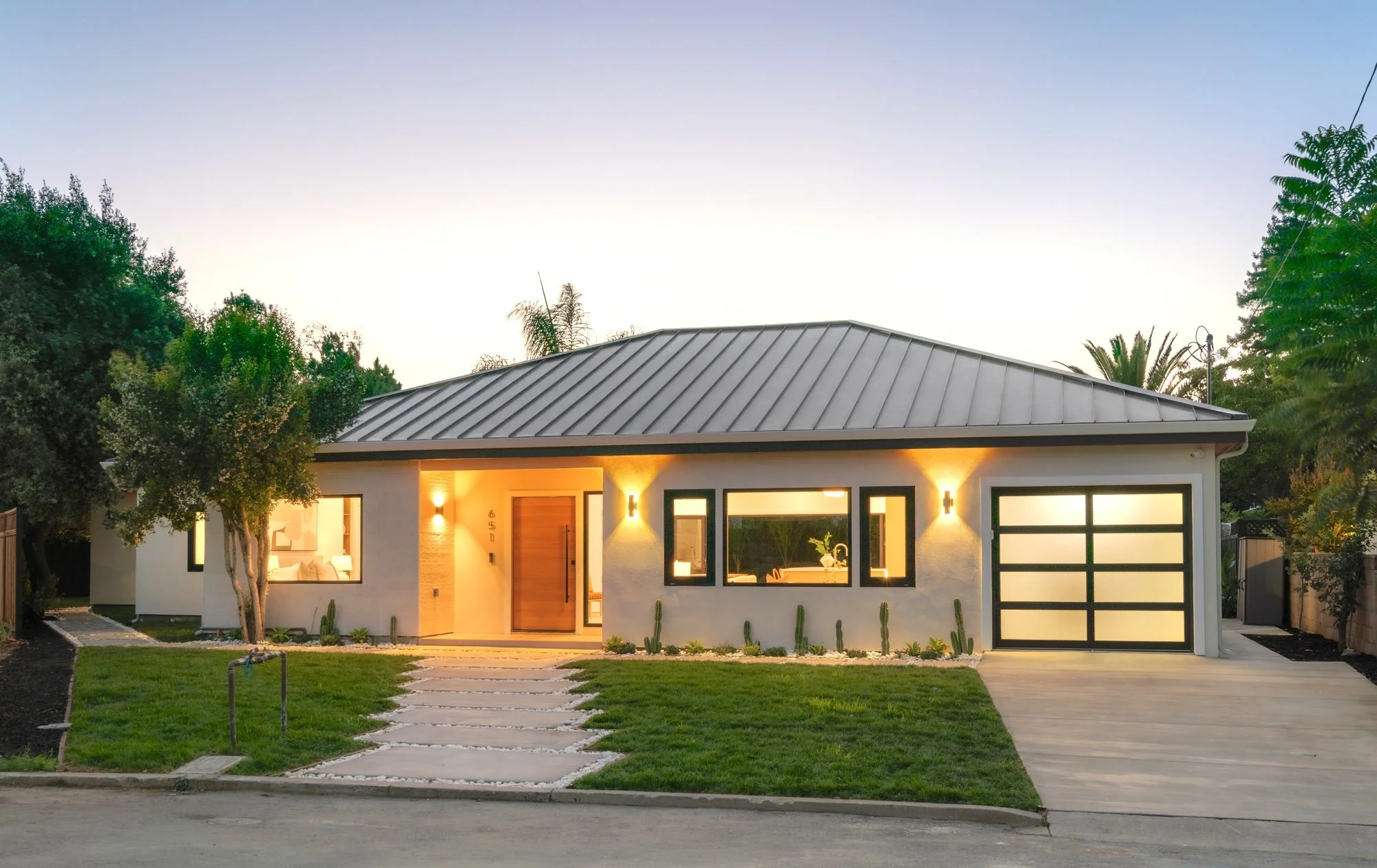 Modern single-story house with large windows and a metal roof, illuminated from inside, front yard with stepping stones and green lawn, trees and clear evening sky in the background.