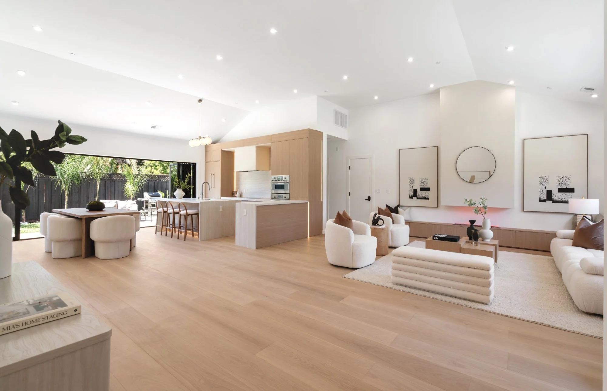 Open-concept living room with light wood flooring, white walls, modern white furniture, and black and white framed artwork. A large sliding glass door leads to an outdoor patio with greenery. Kitchen area features a light wood island with barstools a
