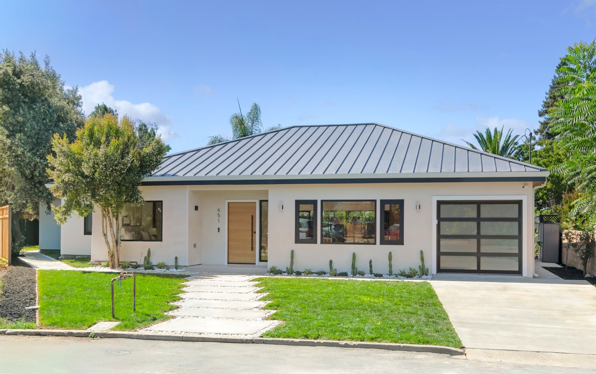 Modern white house with a metal roof, large front windows, and a garage with a glass door. Front yard features a green lawn, pathway, and trees, with a blue sky and clouds above.