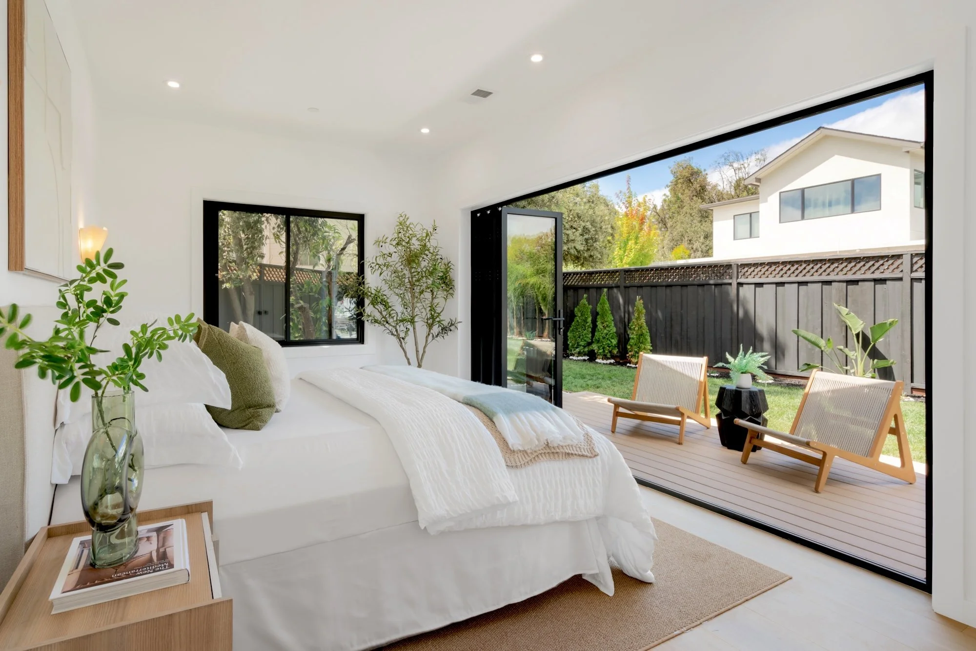 Modern bedroom with large sliding glass doors leading to a backyard with outdoor seating, green plants, and a wooden fence, bright natural light, minimalist decor.