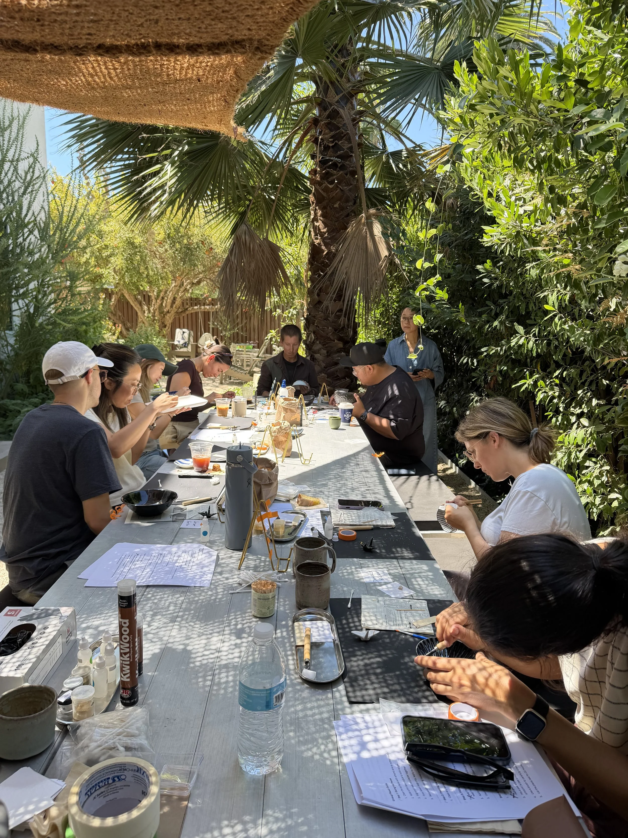 People gathered around a long outdoor table under a palm tree, eating, drinking, and reading papers in a garden setting on a sunny day.