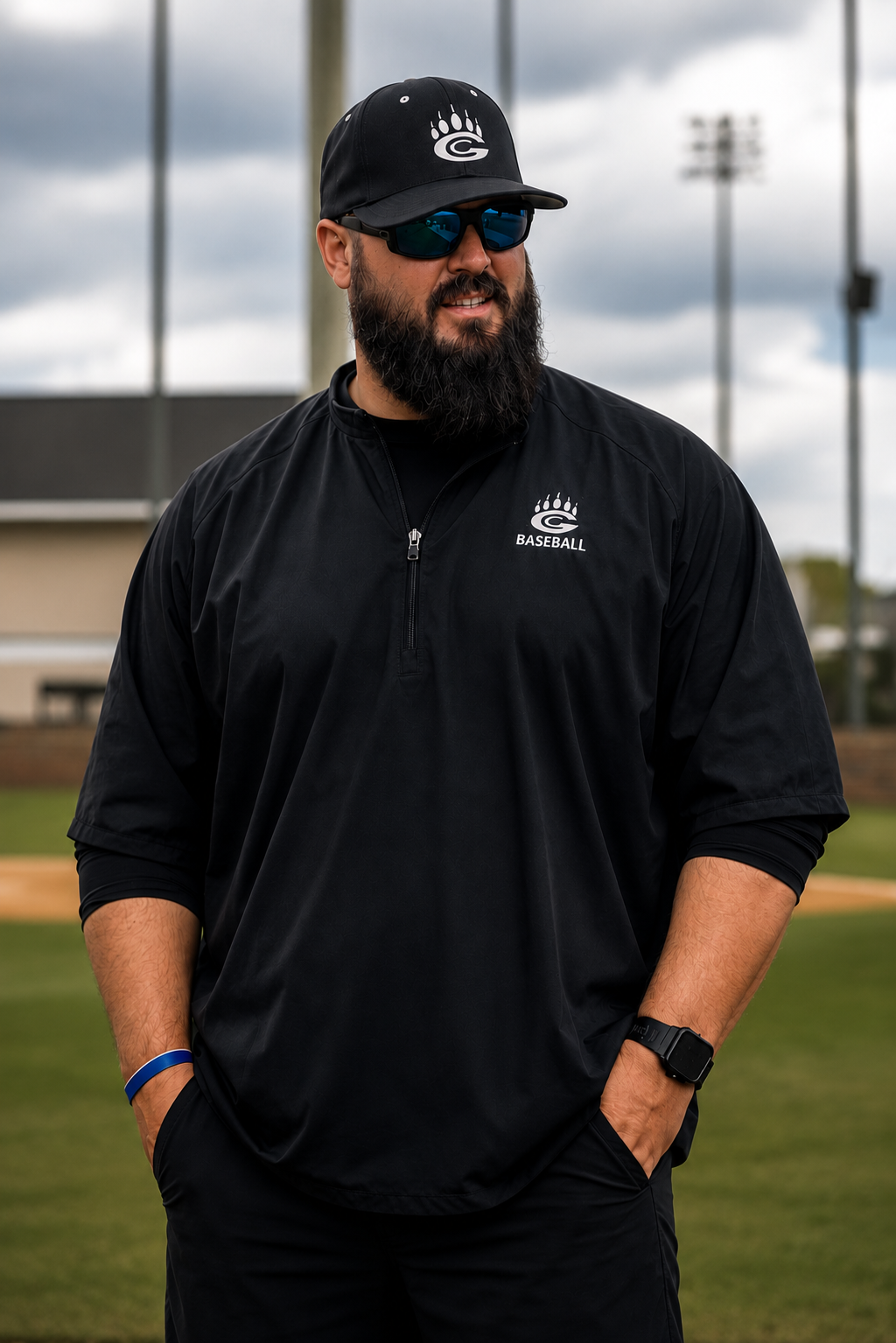 A man with a beard and sunglasses wearing a black Atlanta Braves baseball jacket and cap, standing on a baseball field.