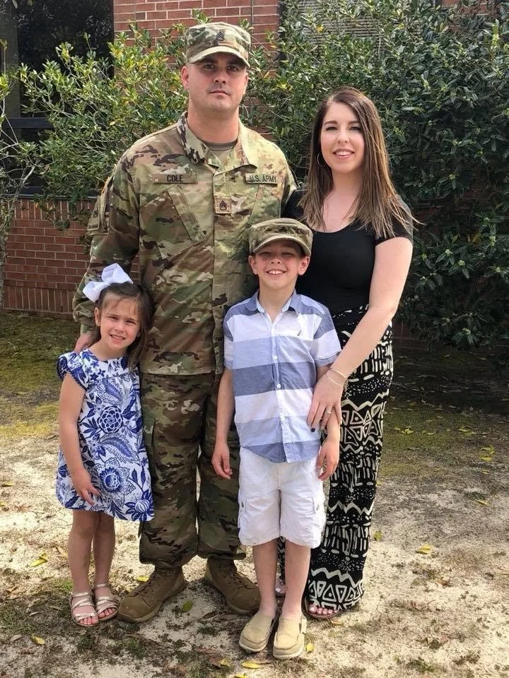 A family of four outdoor, includes a man in military uniform, woman, girl, and boy posing in front of bushes and a brick building.
