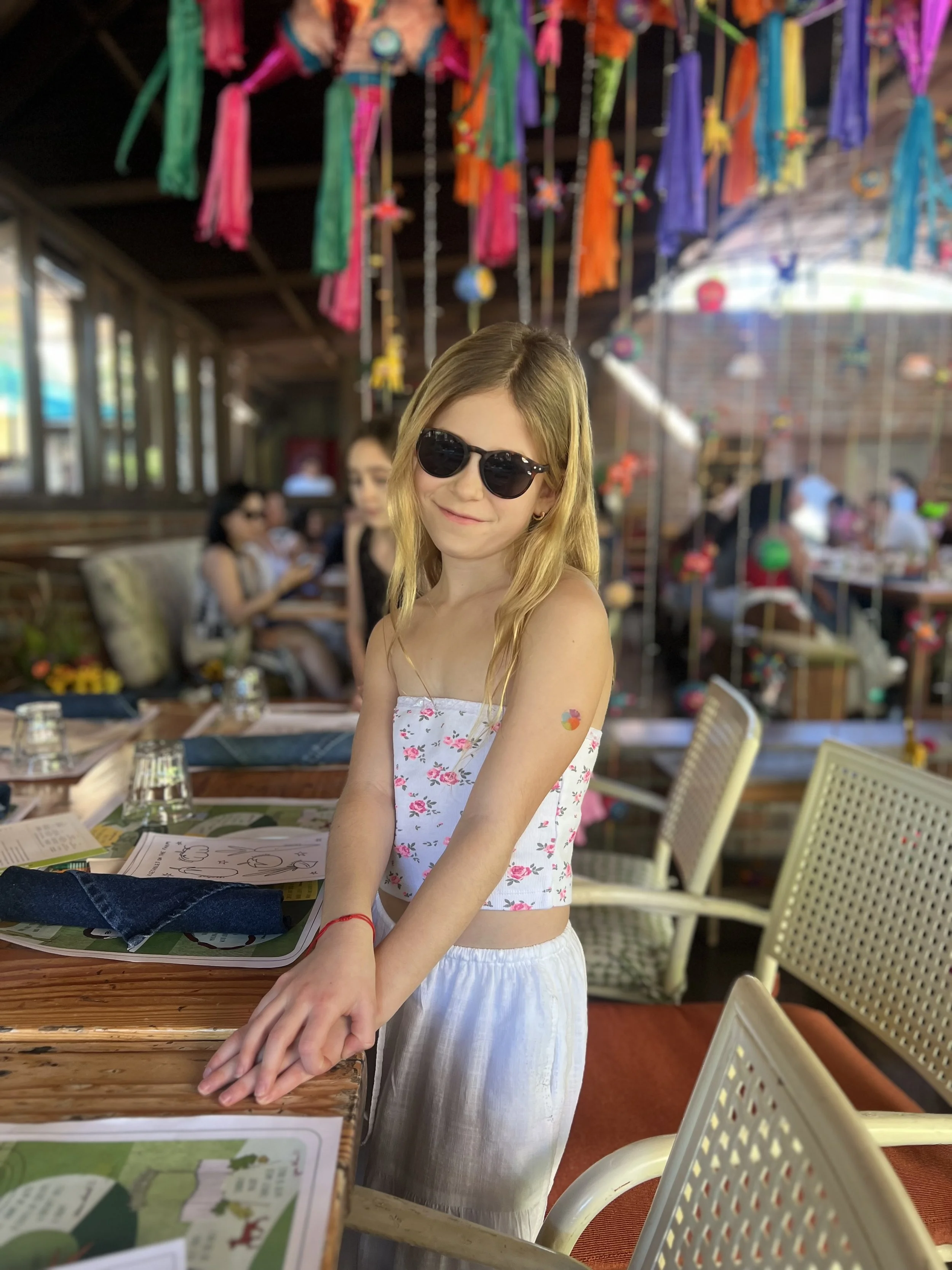 A young woman with blonde hair, wearing sunglasses, a floral strapless top, and white skirt, standing in a decorated outdoor dining area, smiling at the camera with colorful hanging decorations above and other people in the background.