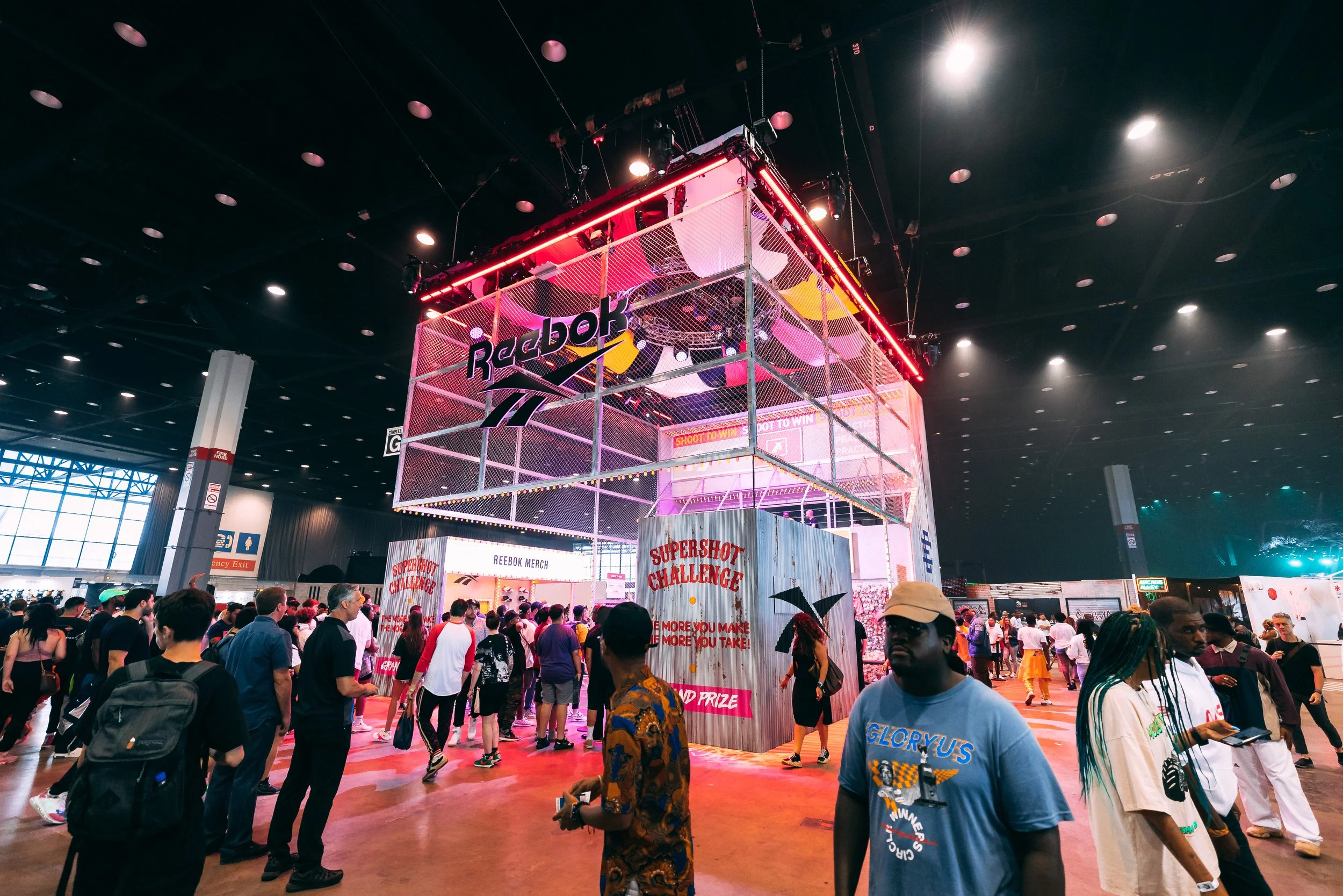 Indoor event with many people gathered around a multilevel Reebok display called SuperShot Challenge, with bright lighting and colorful banners.