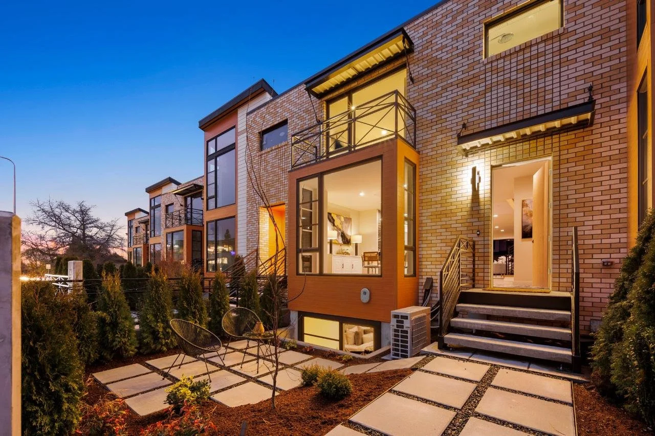 Modern multi-story townhouses with large windows and small outdoor garden patios, during twilight with warm interior lighting.