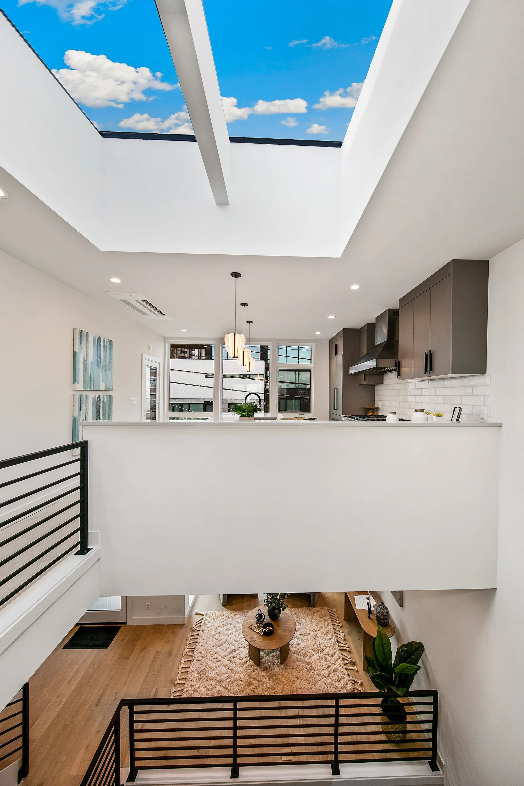 Modern interior view of a two-story home with a kitchen, dining area, and living room. A large skylight shows a blue sky with clouds. The space has white walls, gray cabinets, pendant lighting, and wooden flooring.