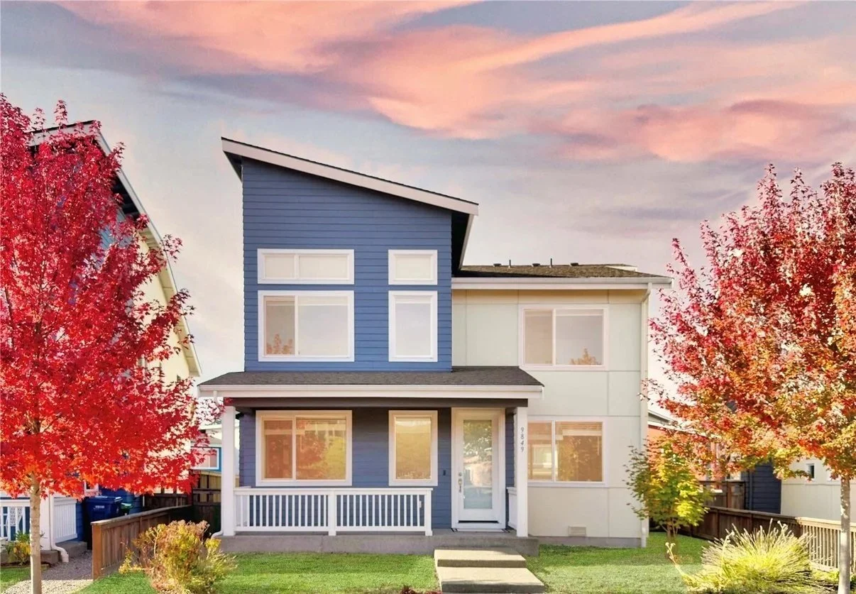 Modern two-story house with blue and white exterior, large windows, and a front porch, surrounded by trees with red autumn leaves at sunset.