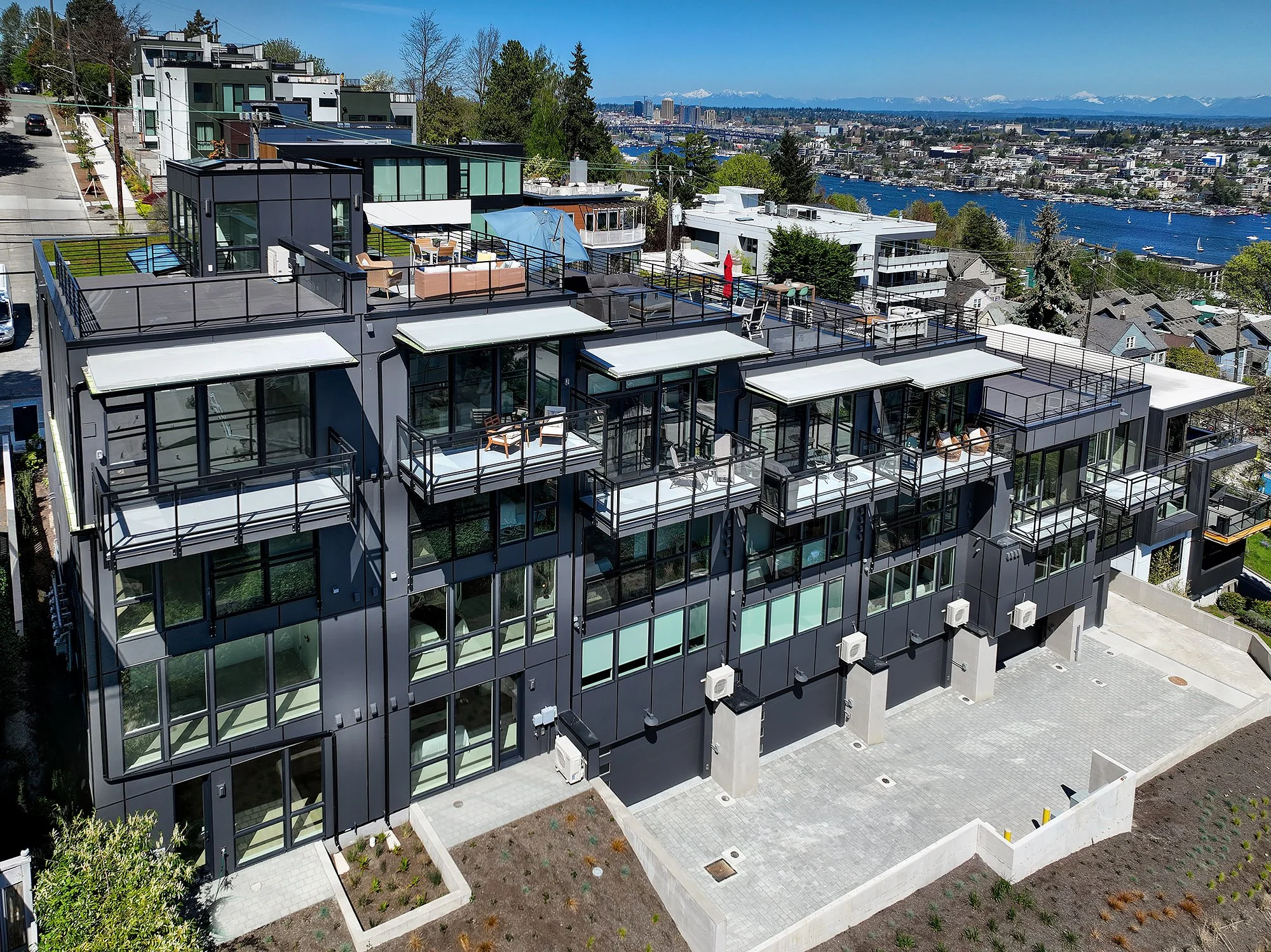 A modern apartment building with glass balconies, some decorated with outdoor furniture, situated on a hillside with a cityscape and water in the background.