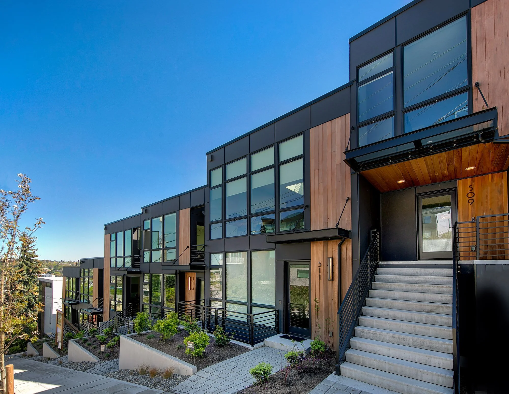 Modern multi-story residential building with large windows, wooden accents, exterior staircase, and landscaped garden area with small plants and trees.