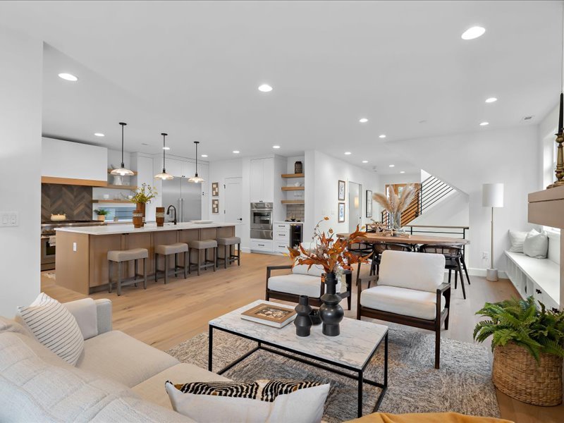 Open-concept modern living room and kitchen with white walls, hardwood flooring, white cabinetry, a kitchen island with bar stools, a dining area, and decorative plants and artwork.