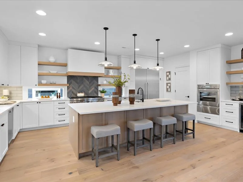 Modern white kitchen with central island, wooden stools, pendant lighting, stainless steel appliances, open shelving, and light wood flooring.