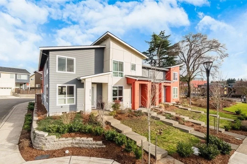 Two-story modern residential buildings with landscaped front yards, concrete pathway stairs, and leafless trees, under a partly cloudy sky.