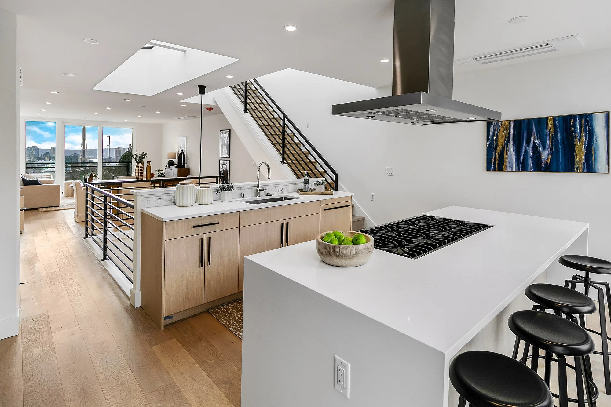 Modern kitchen with white countertops, light wooden cabinets, black barstools, and a city skyline view through large windows.