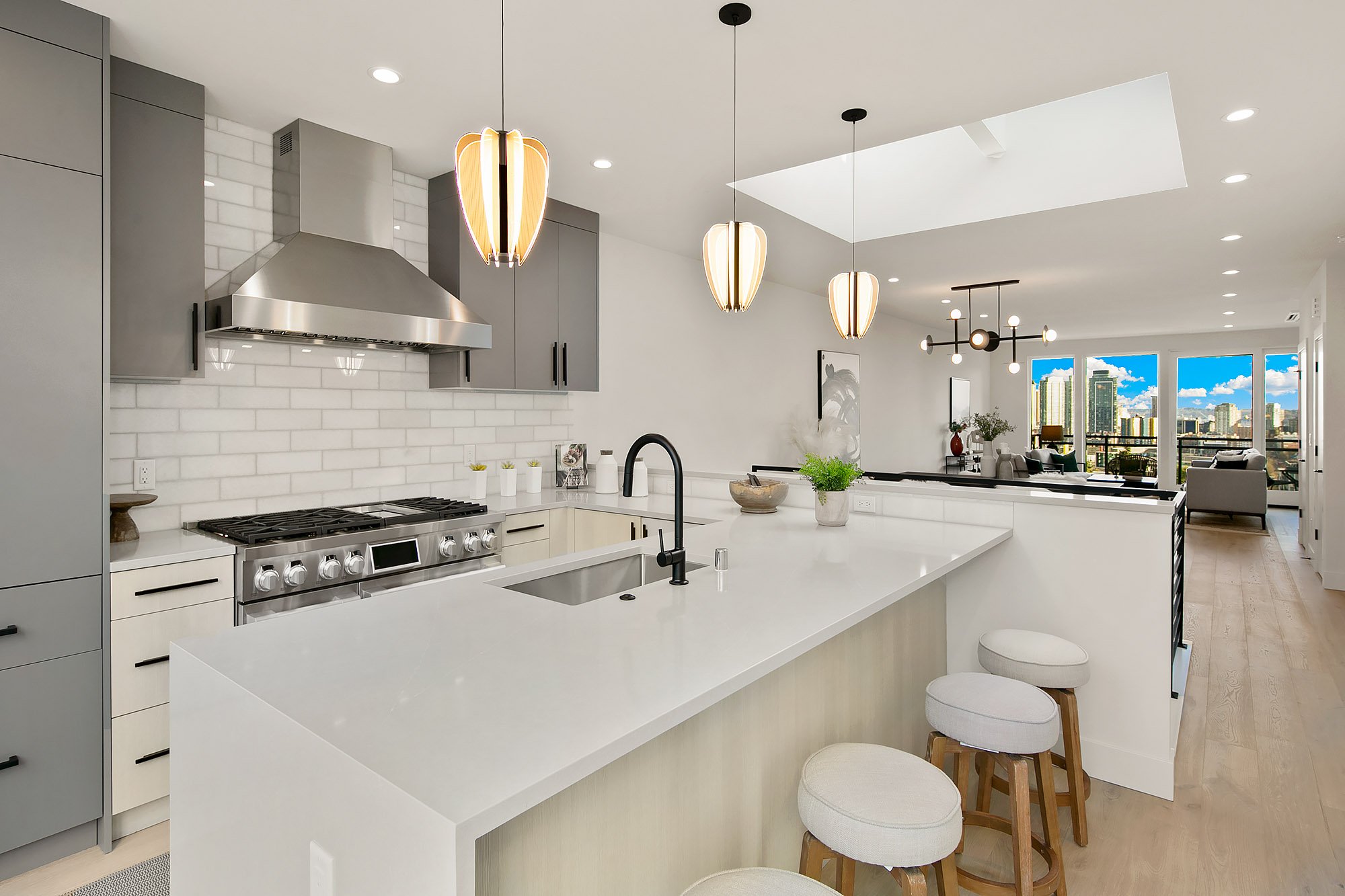 Modern kitchen with white island, gray cabinets, stainless steel stove, white subway tile backsplash, black faucet, hanging pendant lights, open living area with large windows and cityscape view, and natural light from skylight.