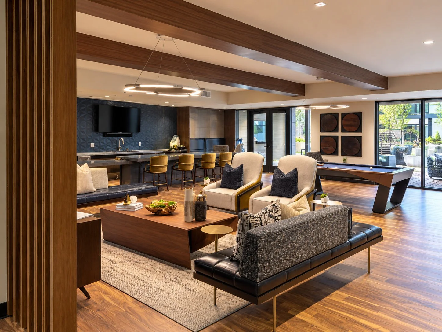 Modern living room with wood flooring, beige armchairs, a black leather sofa, a wooden coffee table, and a pool table, with large glass doors leading outside.