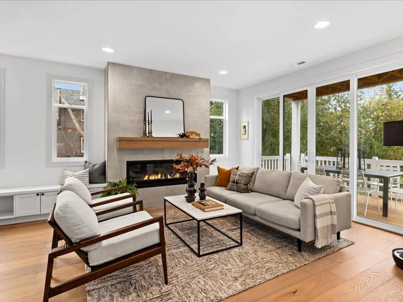 Living room with a beige sofa, two armchairs, a fireplace with a wooden mantel and decor, a rug, and large sliding glass doors leading to a patio with outdoor furniture and trees outside.