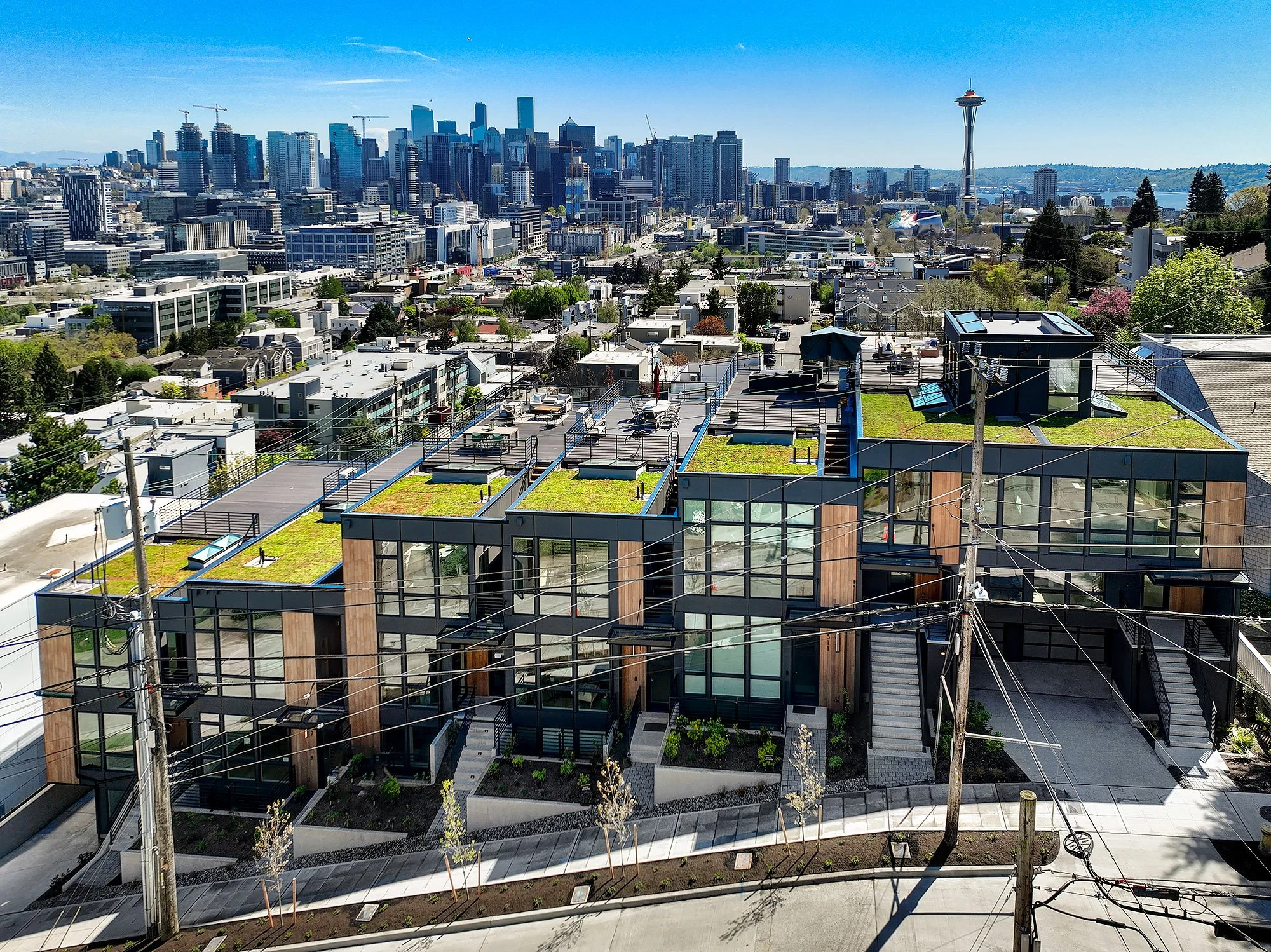 Seattle Residential Development/Seattle skyline with tall buildings and the Space Needle in the background, viewed from a rooftop with modern apartment buildings and trees in the foreground on a clear, sunny day.