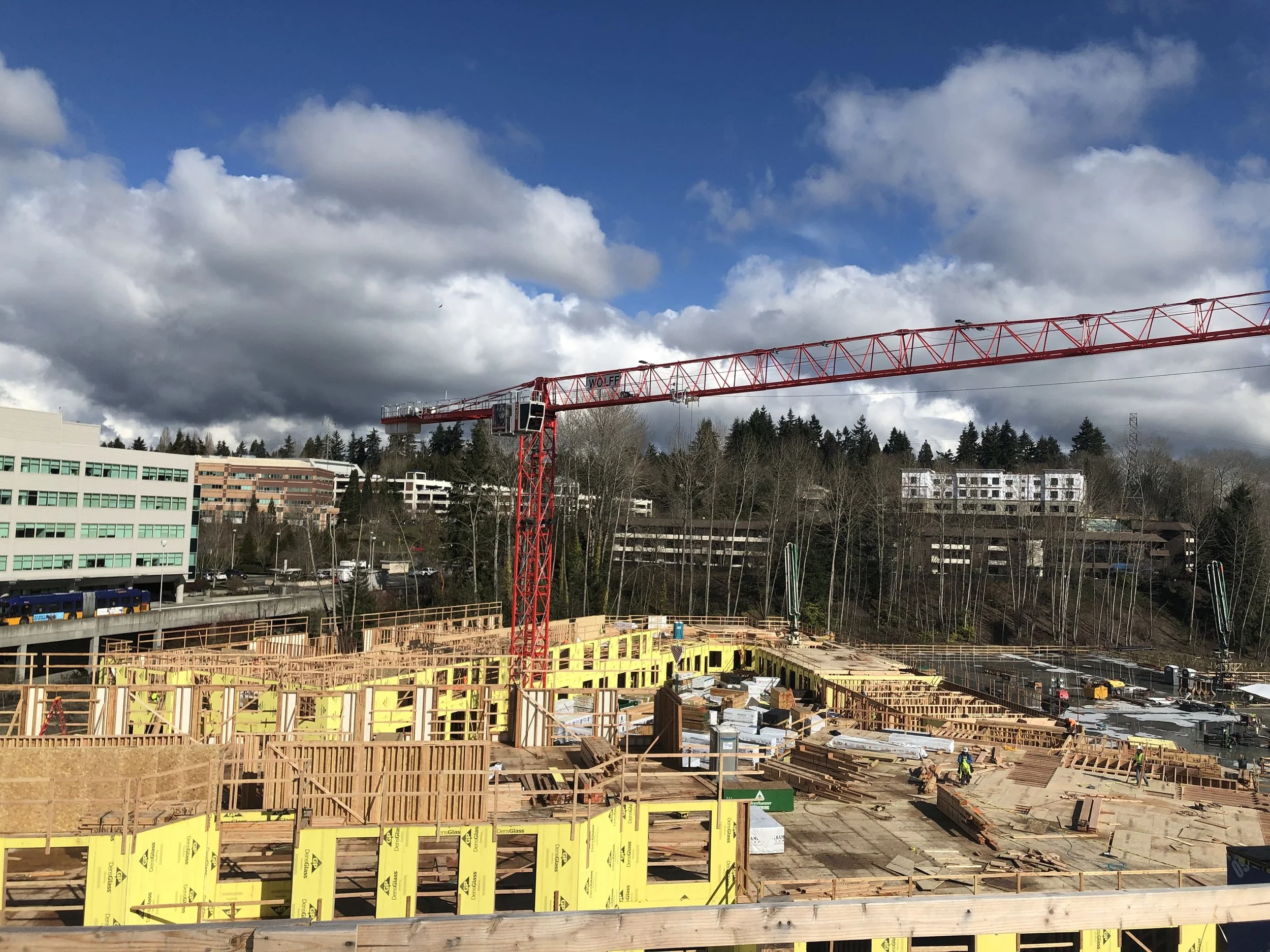 Construction site with wooden framework and a red crane, with buildings, trees, and a partly cloudy sky in the background.
