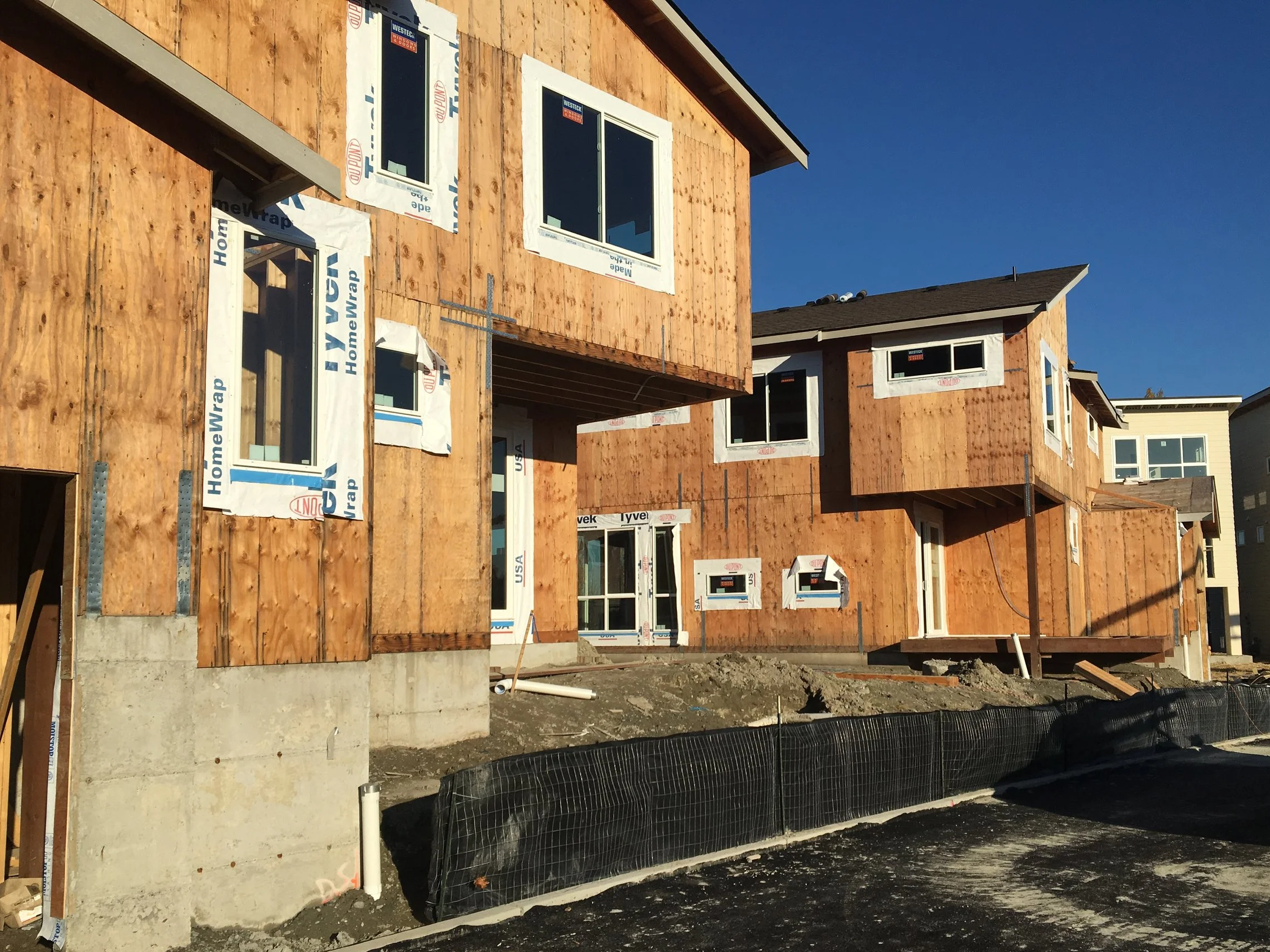 A multi-story house under construction with wooden exterior walls, white-framed windows, and a concrete foundation against a clear blue sky.