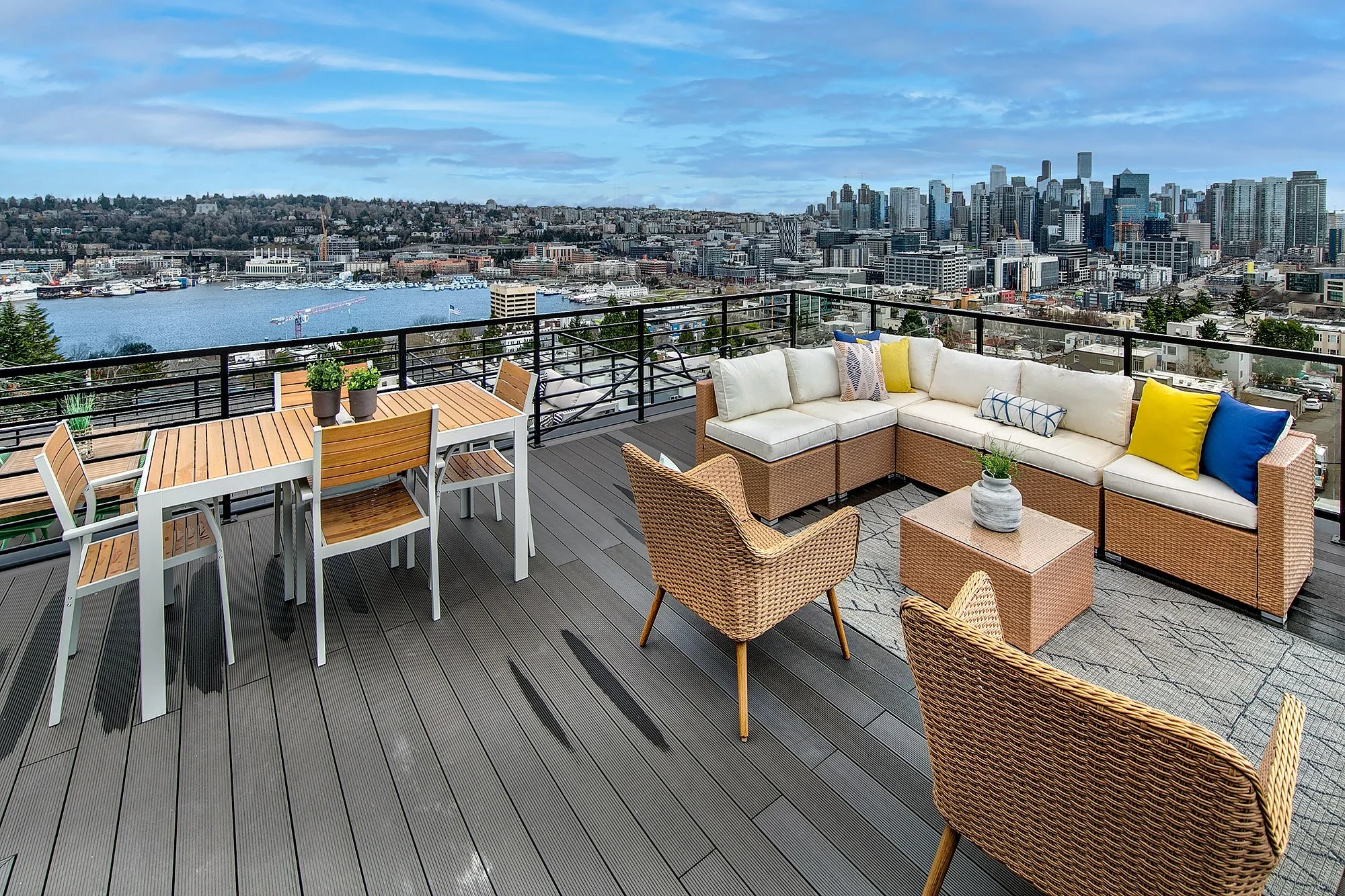 A spacious rooftop balcony with seating and dining area overlooking a city skyline with tall buildings and a harbor.