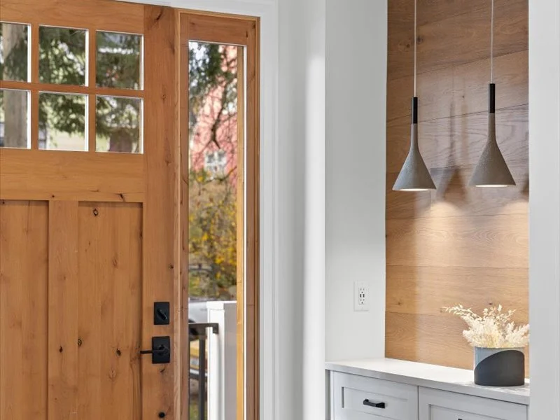Interior view of a modern kitchen or dining area with a wooden front door, a white cabinet, two hanging pendant lights, and a small vase with white flowers on top of the cabinet.