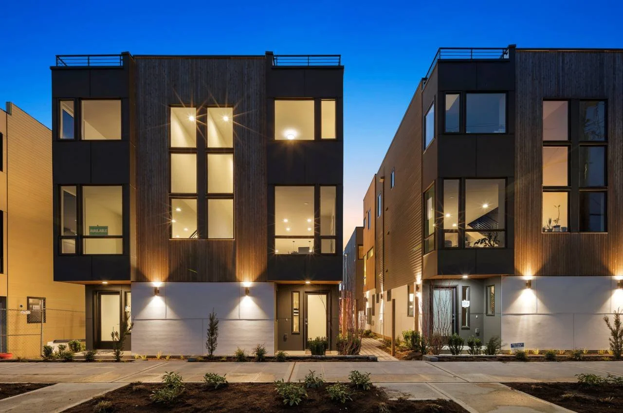 Modern residential buildings with large windows, exterior lighting, and landscaped front yard, during twilight.