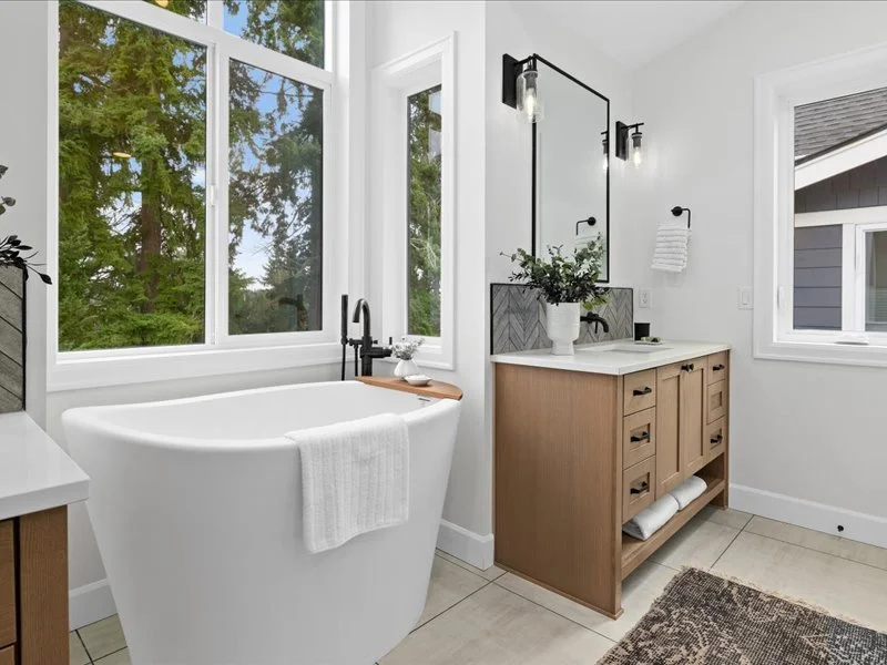 Modern bathroom with a freestanding bathtub near large windows, wooden vanity with a white countertop, framed mirror, and black fixtures, accented by a white towel and lush greenery outside.