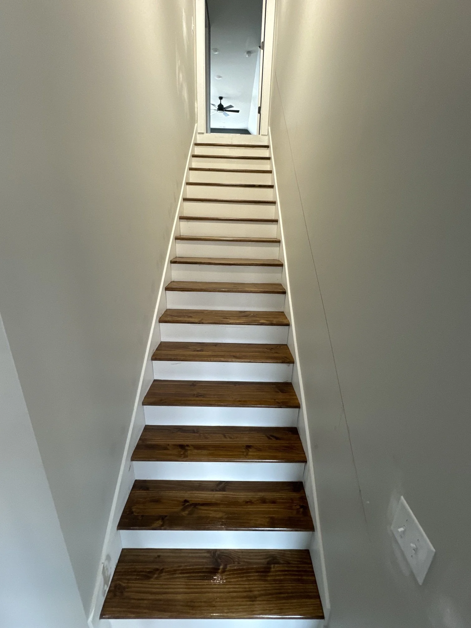 Interior view of a staircase with dark wood steps and white risers, leading up to an open door with a ceiling fan visible in the room beyond.