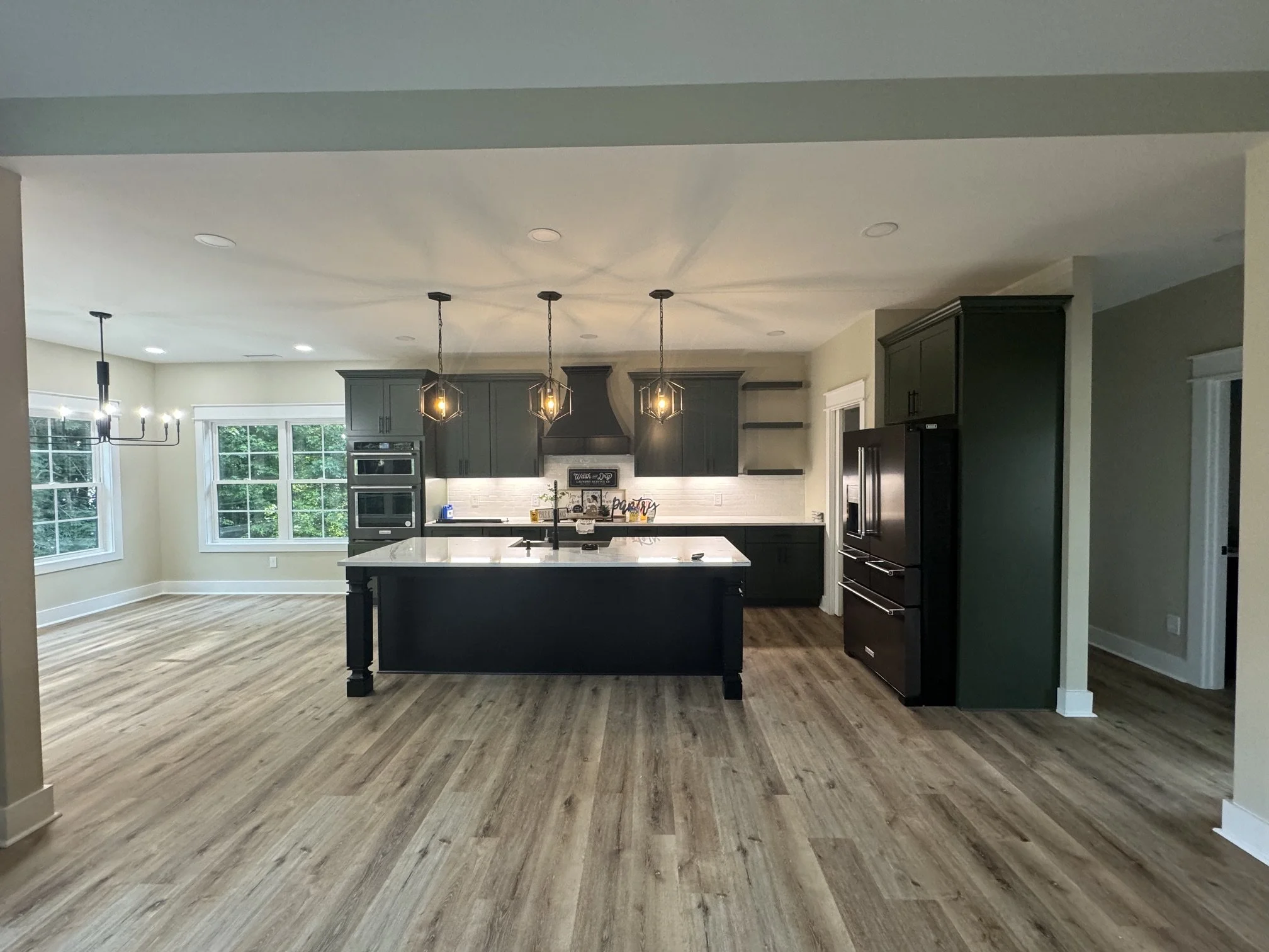 Spacious kitchen with dark cabinets, white island, pendant lighting, and large windows showing greenery outside.