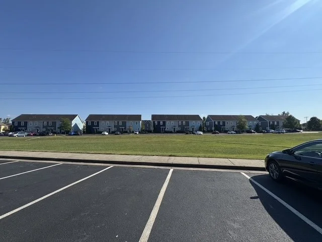An empty parking lot with a grassy area, apartment buildings in the background, and a clear blue sky.