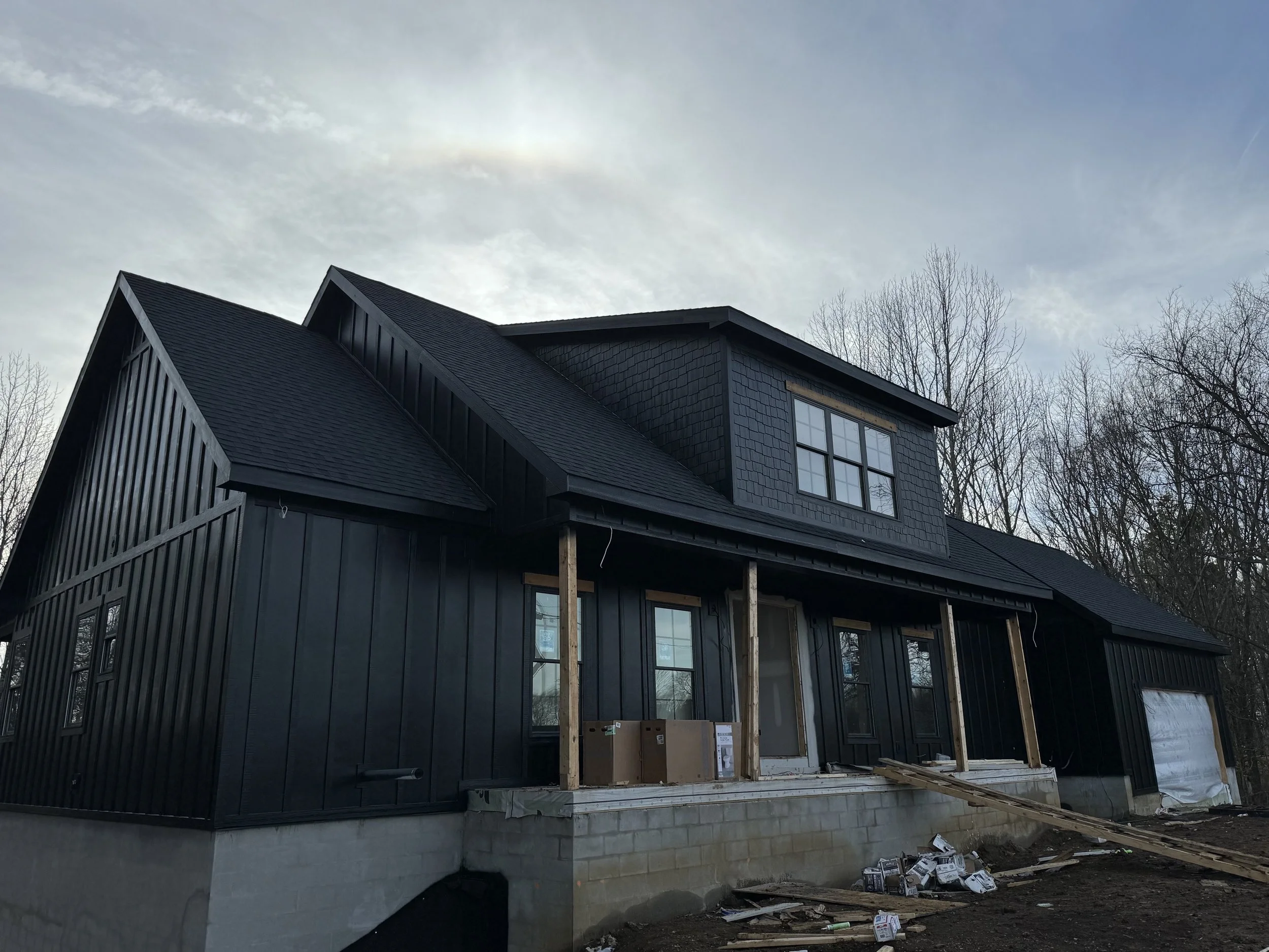 A house under construction with black exterior siding and large windows, surrounded by leafless trees, with construction materials and tools on the ground.