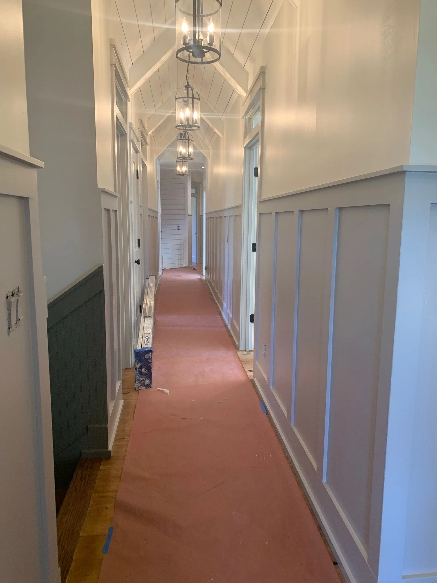 A long hallway with white paneled walls, a vaulted ceiling with white paneling, and a series of hanging light fixtures with glass enclosures. The floor is covered with a pinkish protective sheet, and there are some construction materials and tools along the sides.