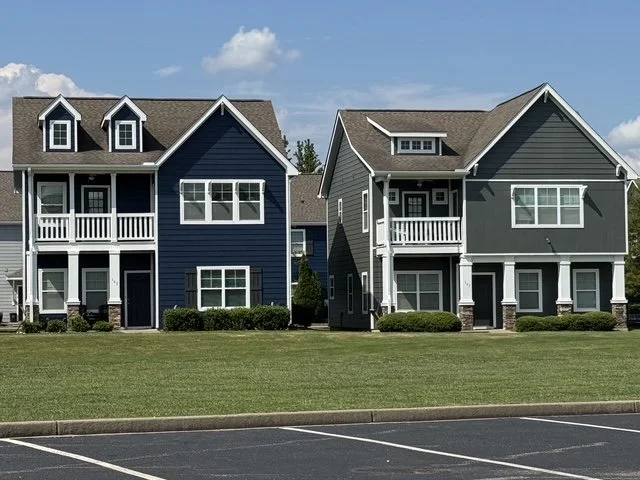 Two modern multi-story houses with front porches, one painted dark blue and the other gray, situated side by side in a residential area with a grassy lawn and parking lot in the foreground.