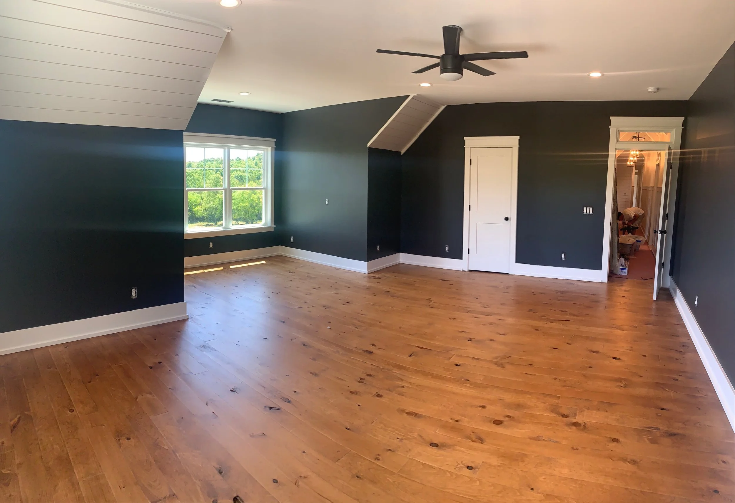Empty room with hardwood floors, dark blue walls, white trim, a window showing greenery outside, a ceiling fan, and a doorway leading to a hallway or another room.
