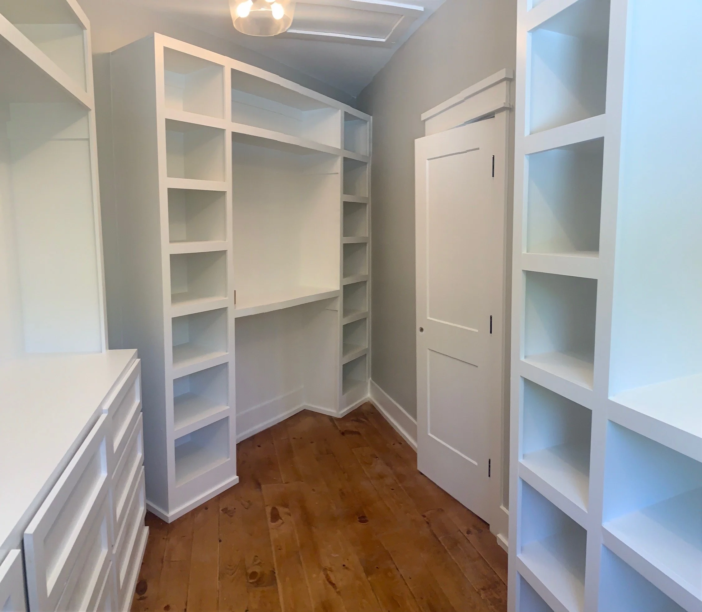 White built-in shelves and cabinets in a walk-in closet with hardwood floors and a ceiling light fixture.