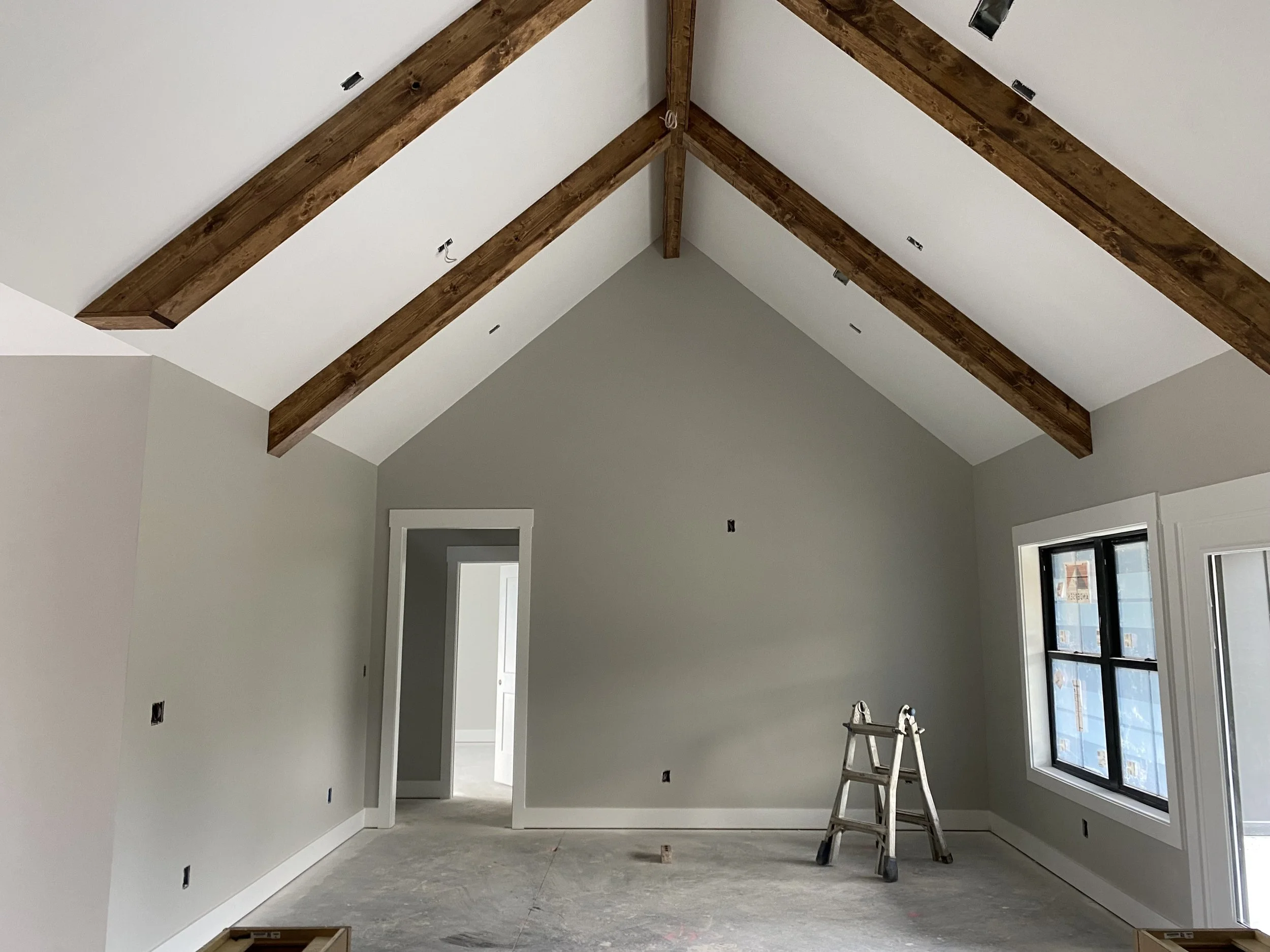 Interior room under construction with exposed wooden beams ceiling, unfinished flooring, a small step ladder, and an open doorway leading to another room with a window.
