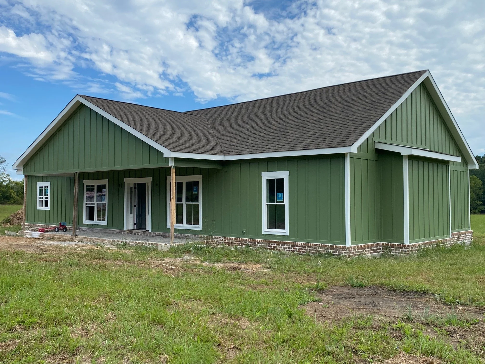 A house under construction with green exterior siding, white trim, multiple windows, and a brown shingle roof. The house has a porch area with support beams, and there is construction equipment and dirt in the foreground. The sky is partly cloudy with patches of blue.