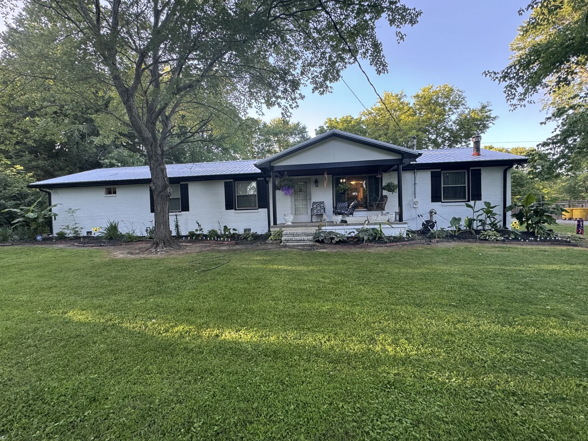 Front view of a white house with black shutters, a porch with outdoor furniture, and a garden with flowers and plants, surrounded by tall trees and greenery under a clear blue sky.