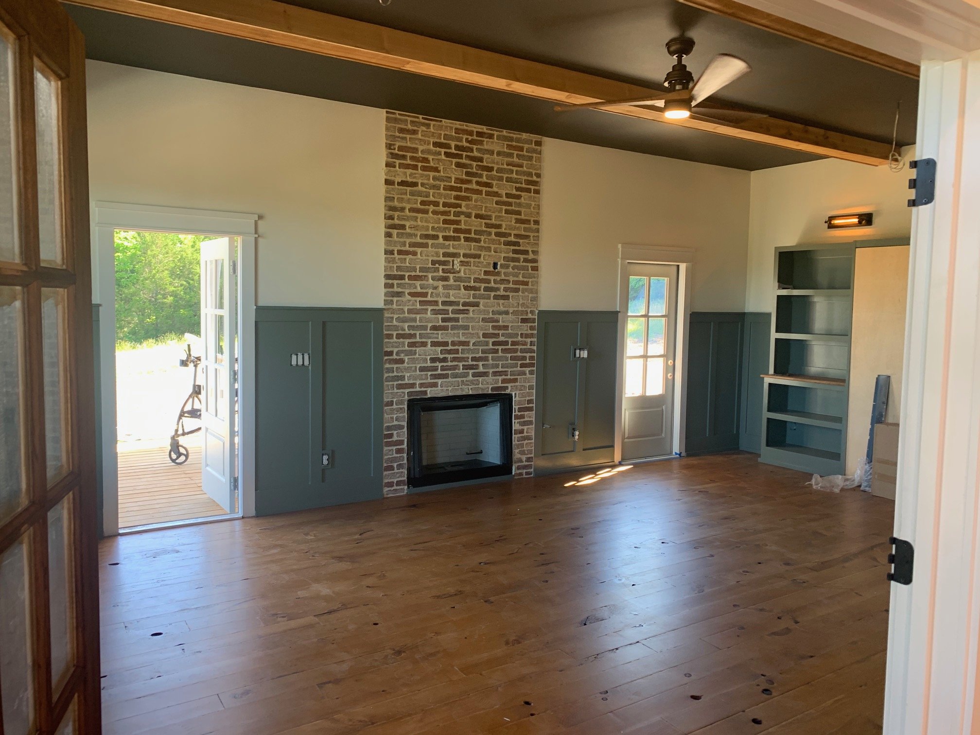 Interior of a room with wooden floors, a brick fireplace, built-in shelves, and large windows letting in natural light. An open door leads outside to a deck.