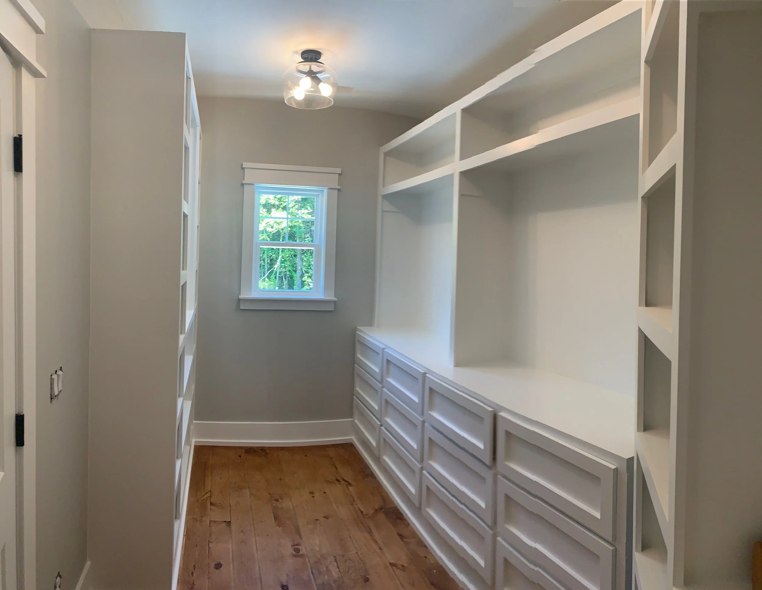 A bedroom closet with white built-in cabinets and shelves, a window showing trees outside, and a ceiling light fixture.