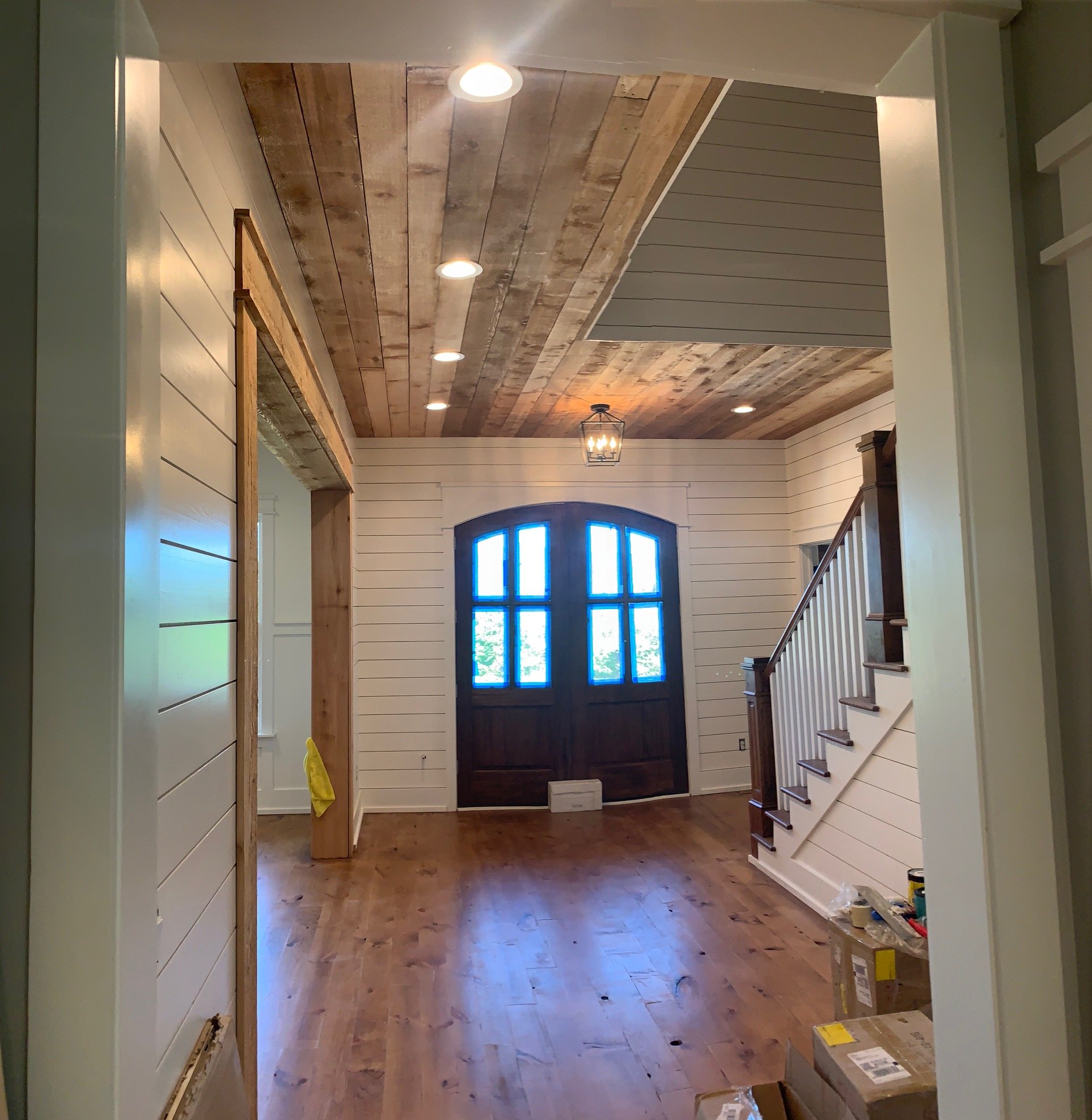 Interior of a house under renovation with wooden flooring, white shiplap walls, a blue arched window, and a ceiling with recessed lighting and a lantern-style light fixture.