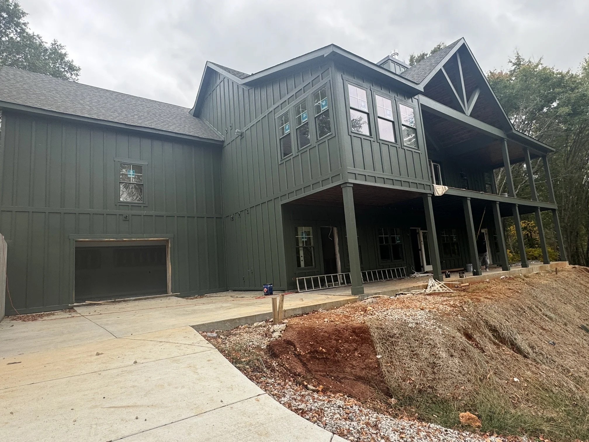 A two-story house under construction with dark gray siding, multiple windows, and a balcony. The construction site has a concrete driveway, dirt, and construction materials around.