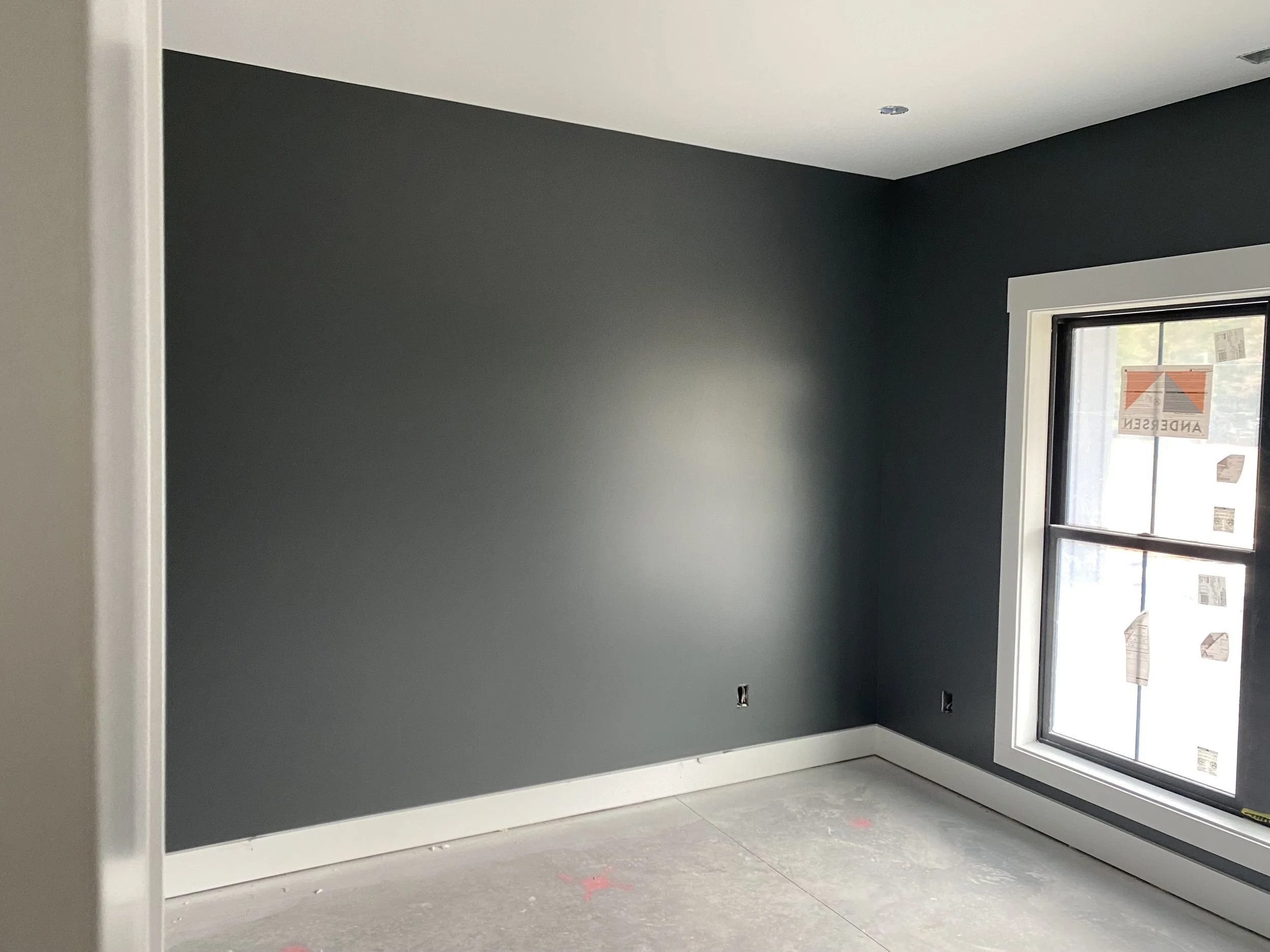 An unfinished room with dark gray walls, a white ceiling, large window with a black frame, and a concrete floor with markings.