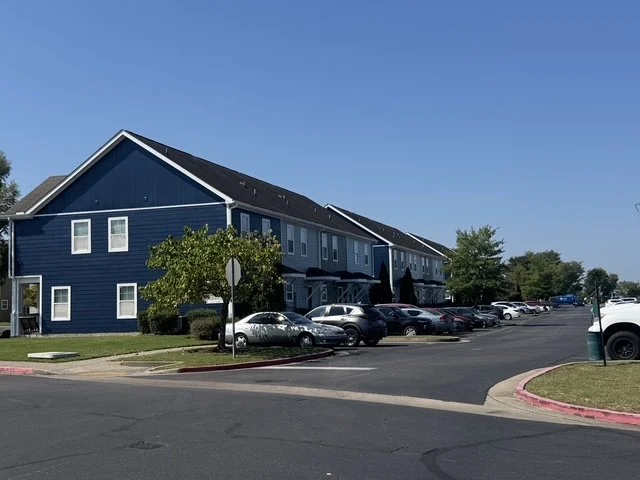 A two-story blue apartment building with a parking lot in front filled with cars, green trees, and a clear blue sky.