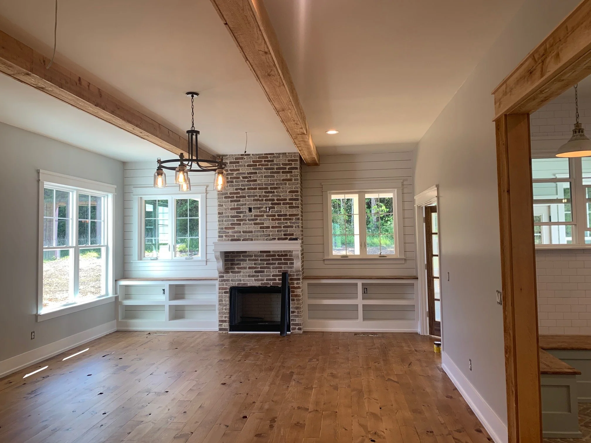 Interior view of a room with hardwood floors, large windows, a brick fireplace, white built-in shelves, and a chandelier hanging from the ceiling.