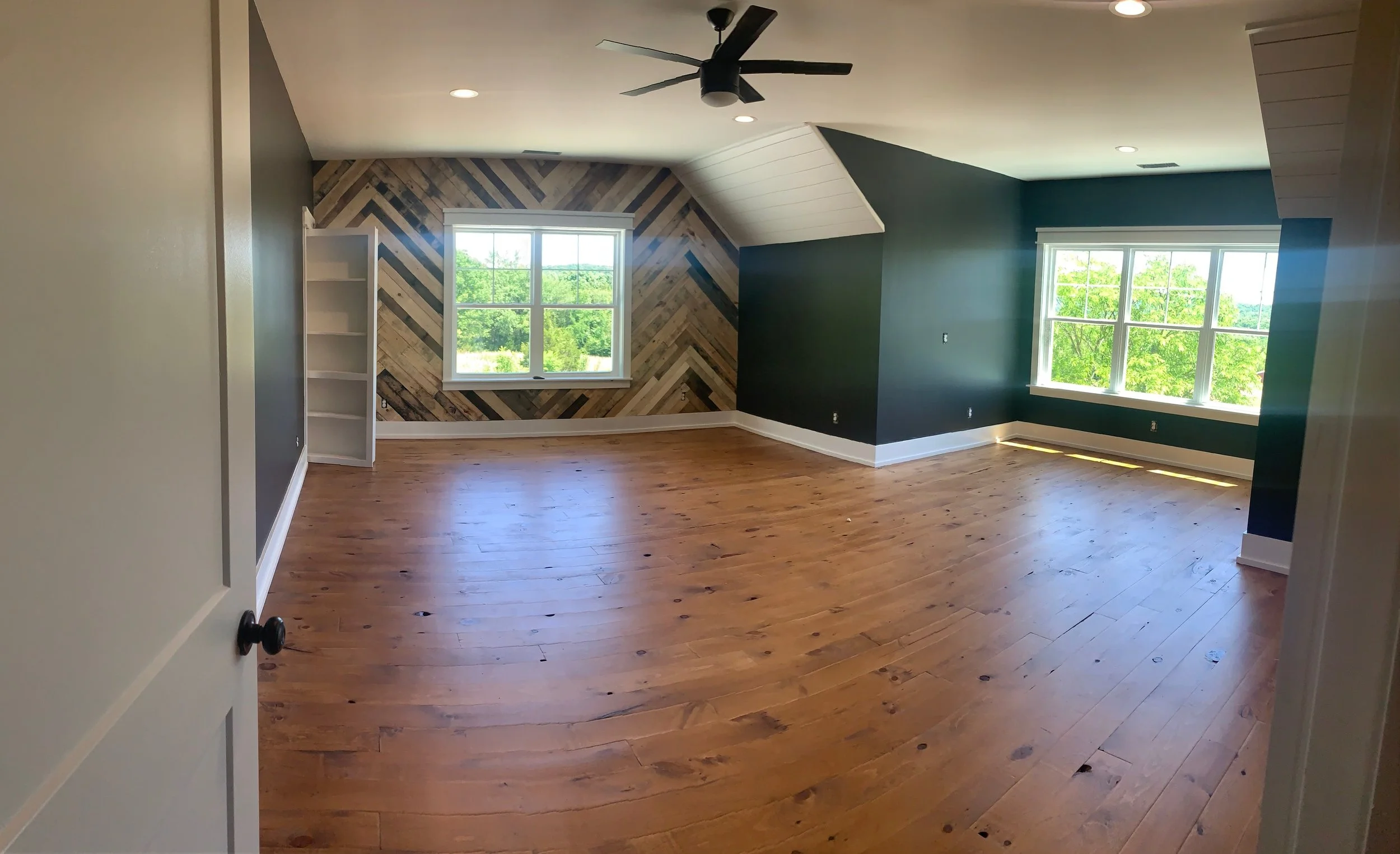 Empty room with hardwood floors, green accent walls, a chevron patterned wood accent wall, large windows with views of green trees, a ceiling fan, and recessed lighting.