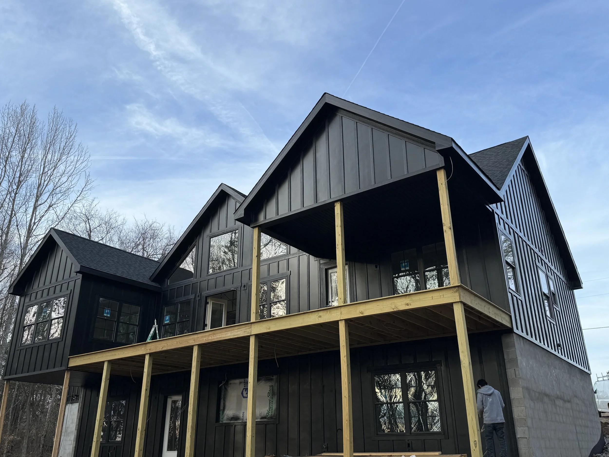 A house under construction with black vertical siding, large windows, and a wooden deck frame. The house has a modern design with multiple gabled roofs, and the sky is mostly clear with some clouds and contrails. Two people are visible near the house, one wearing a gray hoodie and the other a black mask.